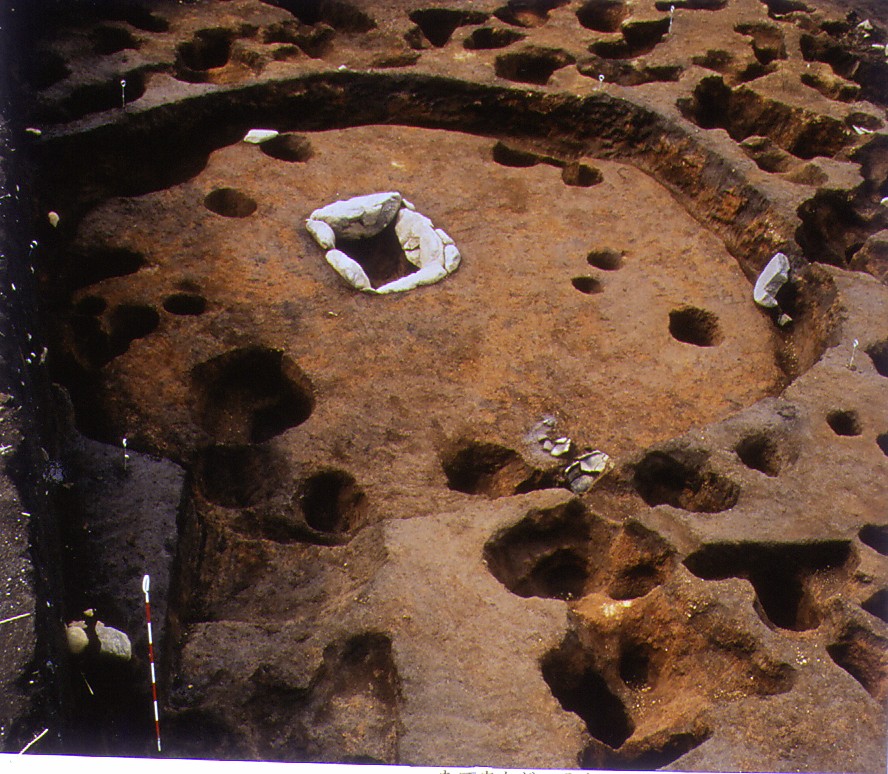 地表から掘り下げられた土の層に円形の穴が多数見られ、中央には石で囲まれた炉跡のようなものがある、遺跡の発掘調査現場の写真