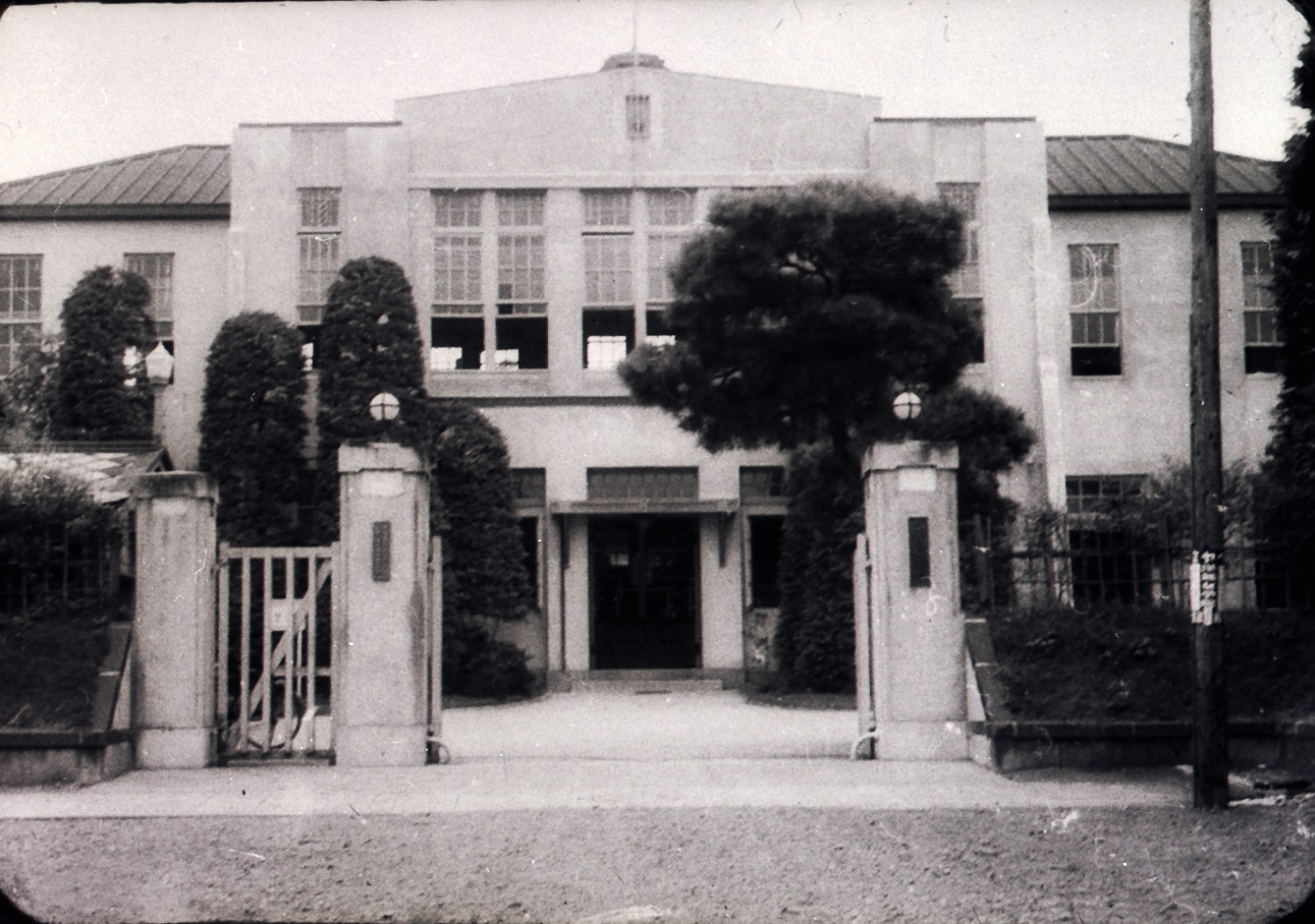 鉄柵と門柱の奥に背の高い木々と洋風の大きな建物が建っている白黒写真