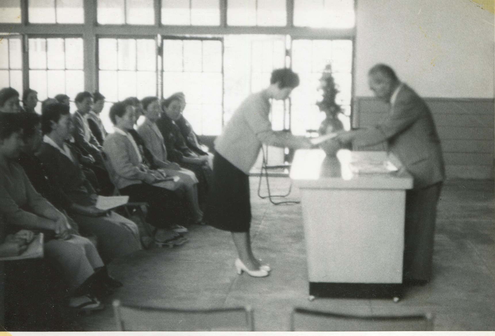 着席したたくさんの人々が見守る中、前に置かれた演台を挟んで男性と女性が書類の受け渡しをしている場面の白黒写真