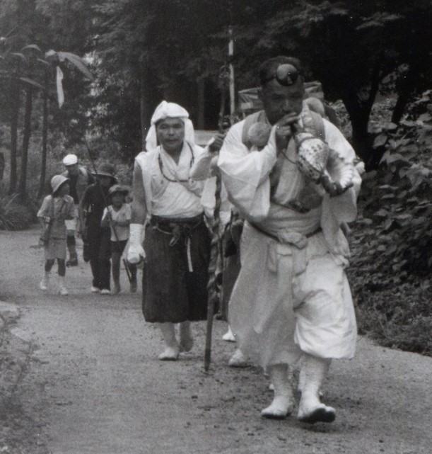 白い山伏の装束を着てほら貝を吹きながら歩く男性を先頭に、大人や子どもが列になって歩いている様子のモノクロ写真