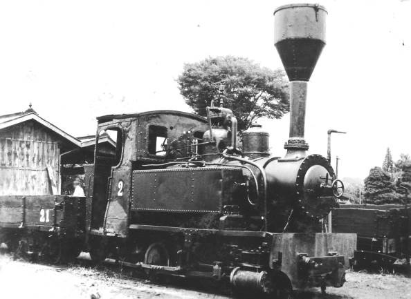 貨車が連結された、大きな煙突の蒸気機関車が駅舎の前に停まっているモノクロ写真