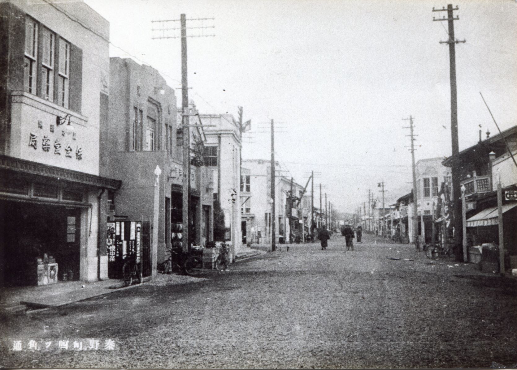 電柱と建物が並ぶ舗装されていない道で、遠くに歩いている人影が見える、昔の街並みのモノクロ写真