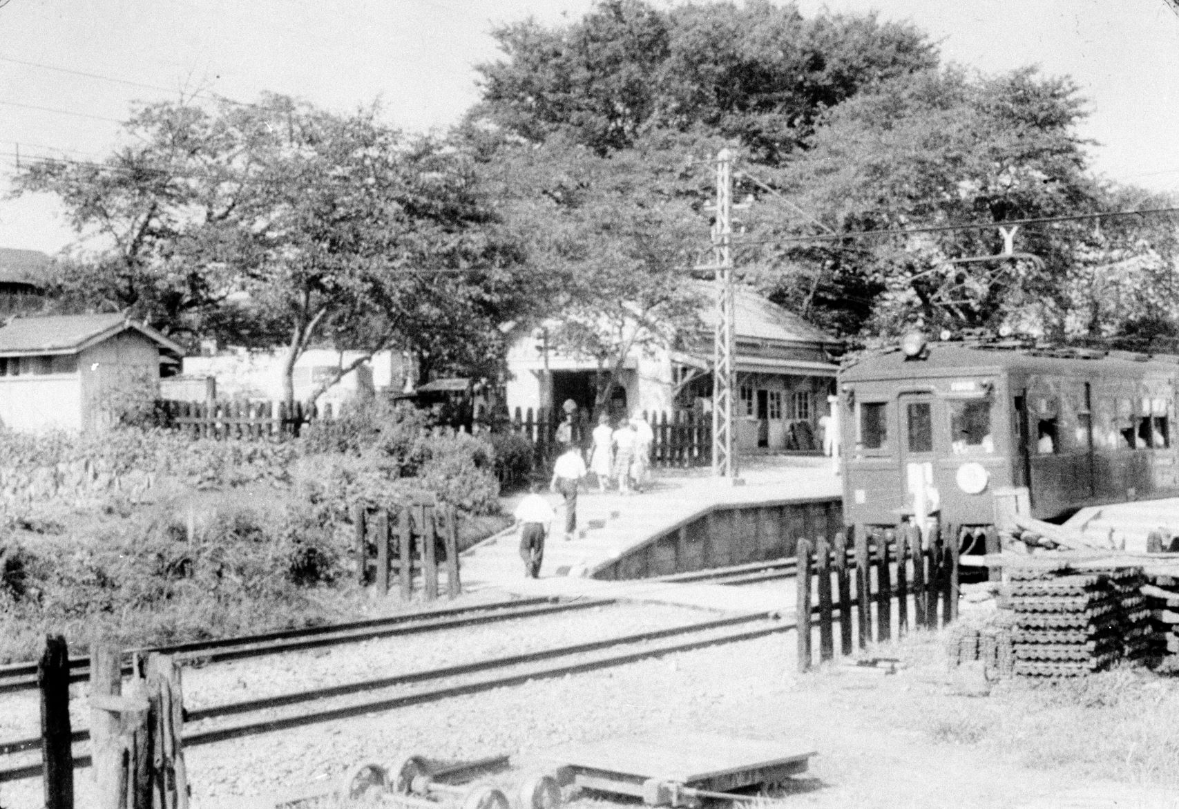 木造の小さな駅舎の前に電車が停まり、複数の人々が駅舎への道を歩いている様子のモノクロ写真