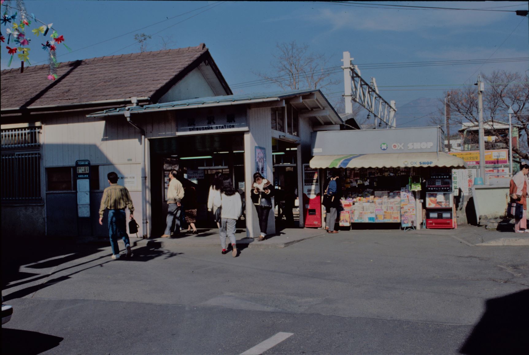 古い駅舎の入り口前を数人の人々が歩いており、駅の隣には小さな売店がある駅前の写真