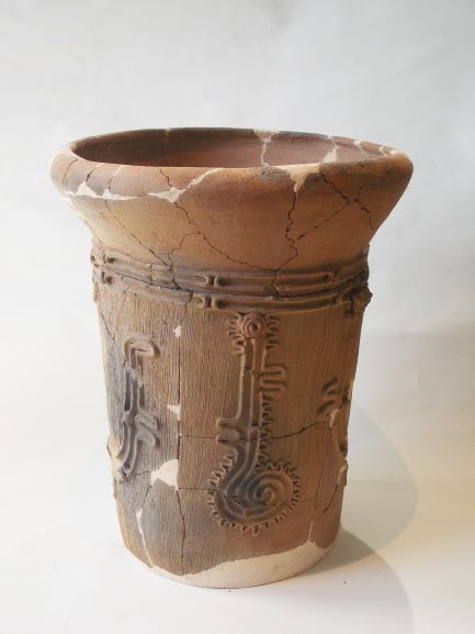 上部が大きく欠けており、全体にひび割れが見られる筒状の土器で、器の胴体部分には複雑な螺旋や波線、渦巻きなどの縄文模様が浮き彫りになって描かれている写真