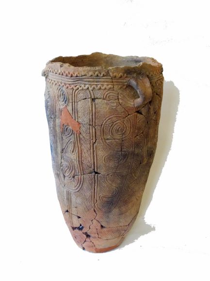 全体がひび割れており、器の表面には渦巻き模様や波線が彫り込まれ、上部に取っ手のような突起がついた、欠片がいくつか剥がれ落ちた状態の土器の写真