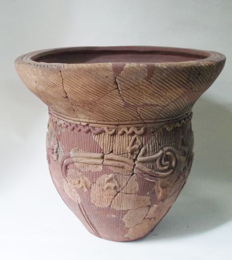 大きく開いた口と丸い胴体が特徴的な土器で、器の表面は全体的にひび割れ、渦巻きや波線のような複雑な縄目模様が彫り込まれている写真