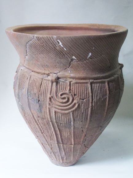 底が尖り、全体にひび割れや欠けが見られる土器で、上部と胴体に縦の線や渦巻き、波線などの模様が施されている写真
