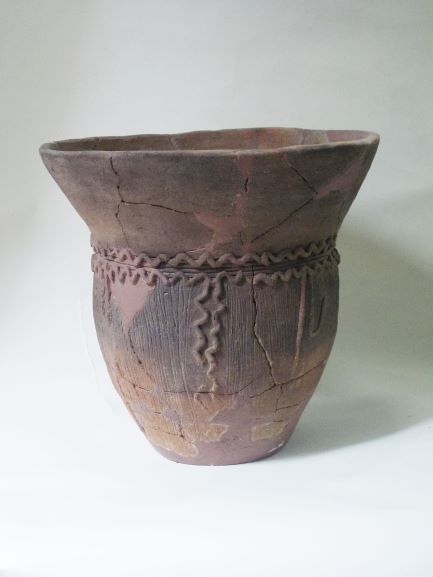 底から上部にかけて広がる形の土器で、器の表面にはひび割れや欠けが見られ、渦巻きや波線のような複雑な文様が施されている写真