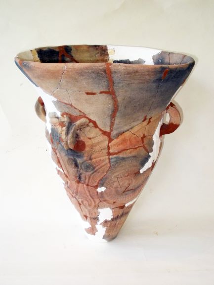 逆三角形のような形状をしており、全体的にひび割れや欠けが見られ、渦巻きや波線のような複雑な文様が施されている土器の写真