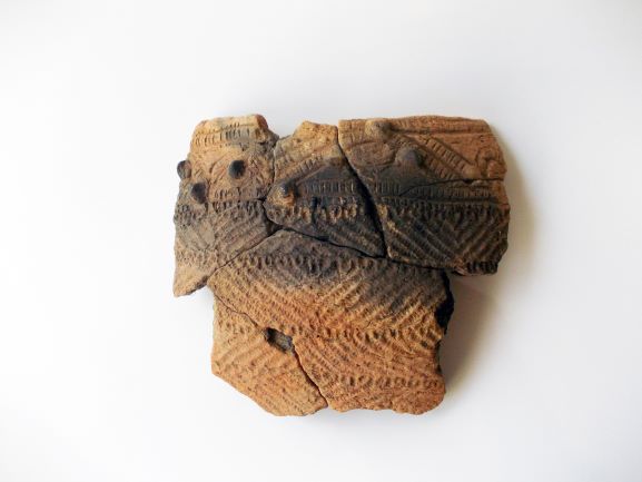 複数の破片を組み合わせて修復された土器の一部で、表面にはいくつかの文様や隆起した装飾が施されている写真