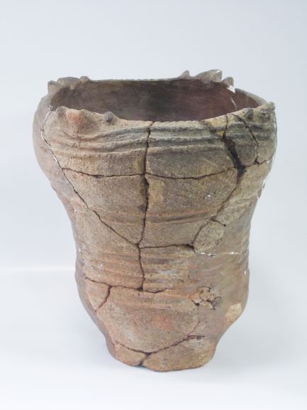 いくつかの破片をつなぎ合わせて修復された、上部にふくらみがある深鉢形で、表面に横方向の筋のような文様が刻まれている土器の写真