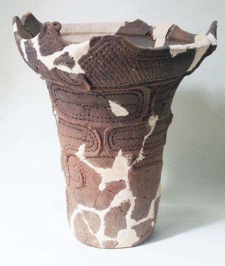 白い補修材でつなぎ合わされた、深鉢形で、表面には様々な模様が施され、上部の口縁部分が波打つように作られている土器の写真