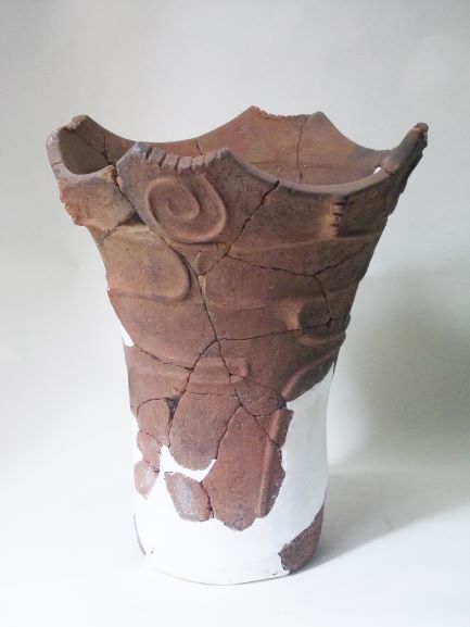 白い補修材でつなぎ合わされた、深鉢形で、表面には渦巻きのような模様が施され、上部の口縁部分が波打つように作られている土器の写真