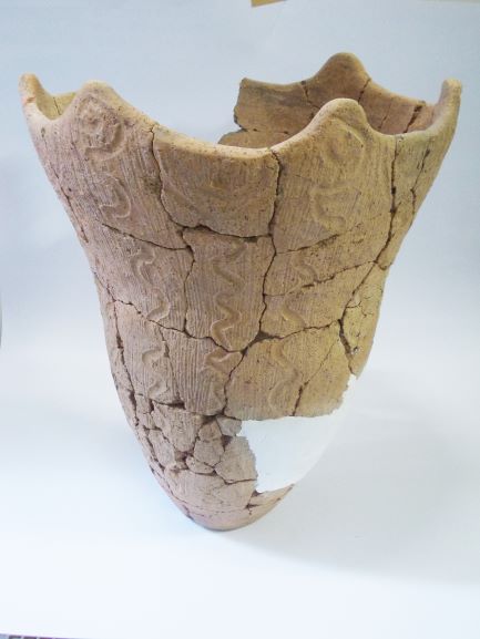 一部が白い補修材でつなぎ合わされた、深鉢形で、表面には縦に波型の模様が施されている土器の写真