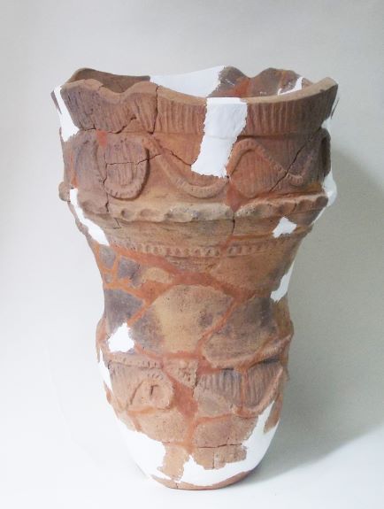 白い補修材でつなぎ合わされた、深鉢形で、表面に波や直線の模様が複雑に施され、上部の口縁部分が波打つように作られている土器の写真