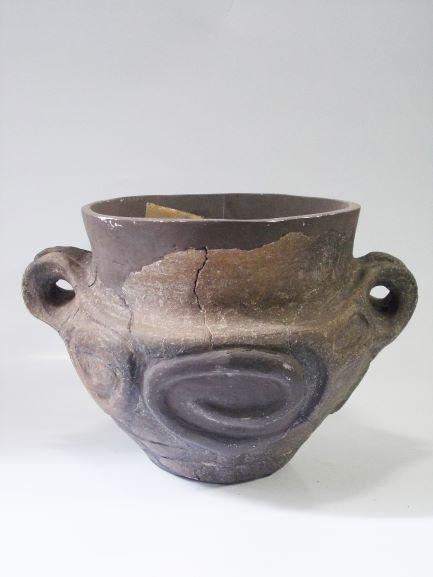 薄茶色と深茶色の鉢型で、左右に持ち手があり、立体的な渦巻き模様の装飾がされた土器の写真