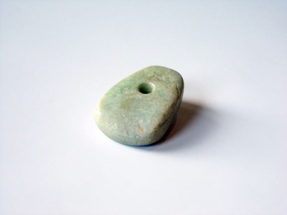 薄い緑がかった色で、中央付近に小さな丸い穴が開けられている楕円形の石の写真