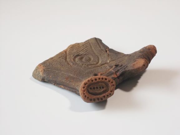 複数の円形の突起と渦巻き模様が組み合わされた複雑な文様が施されており、持ち手のようなものがある土器の写真