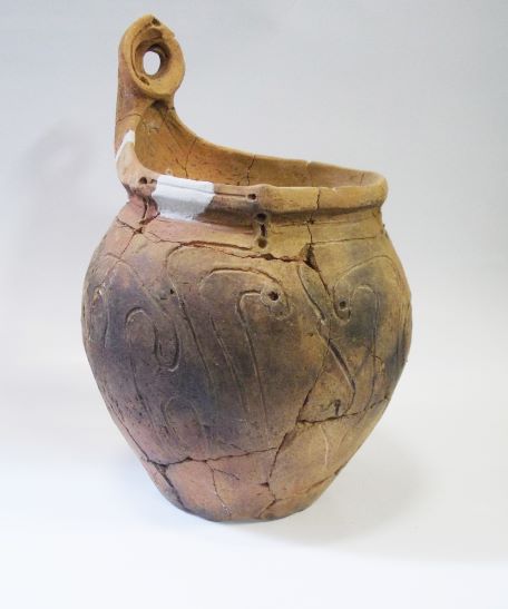 複数の破片を組み合わせて修復された深鉢形で、表面には模様が施され、持ち手のような突起がついている土器の写真