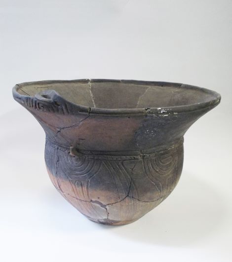 球状の胴体から口縁部にかけて大きく広がった鉢形で、胴部には複数の渦巻き文様が施され、口縁部には一つの突起が確認できる土器の写真