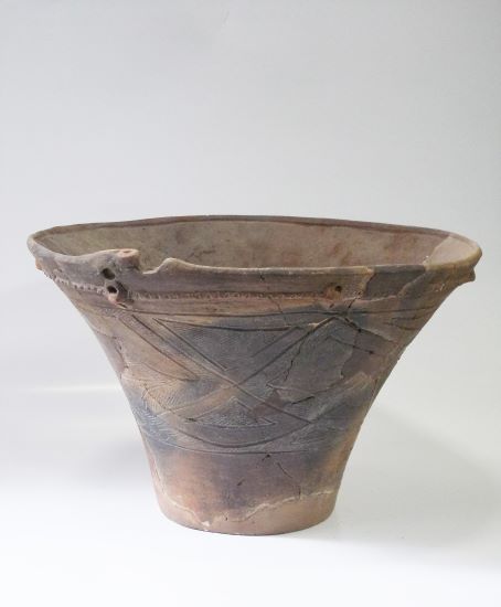 口縁部が大きくラッパ状に広がり、胴部には複数の三角形や斜め線で構成された文様が施され、ひび割れや変色が見られる深鉢形土器の写真