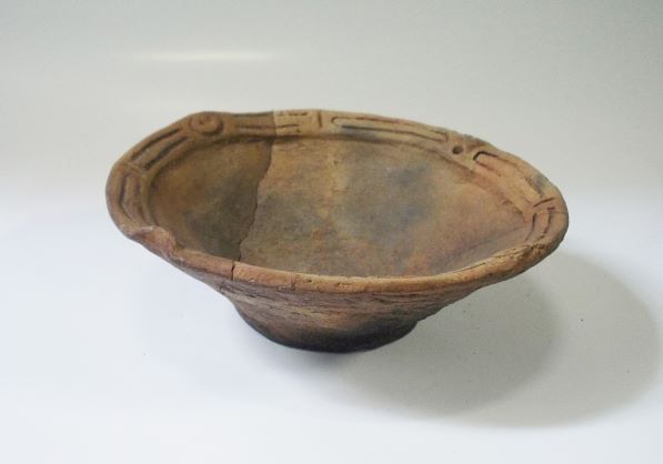 大きく開いた口縁部から底部にかけて浅く広がった浅鉢形で、口縁部の表面には文様が施されており、全体的にくすんだ茶色をした土器の写真