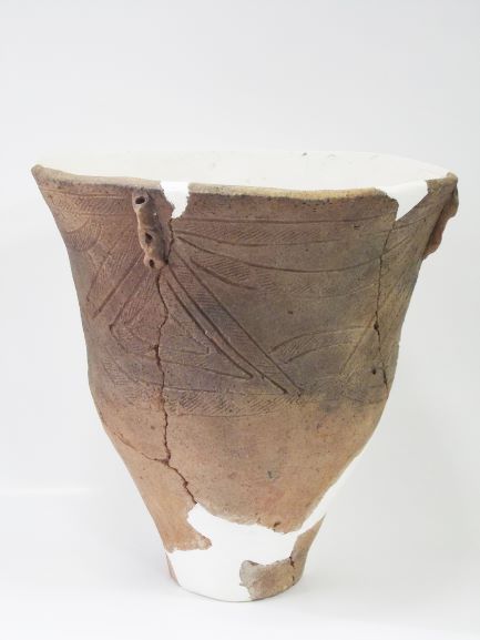 口縁部が大きく広がり、胴体には植物の葉のような文様が施され、ひび割れや一部が白い修復跡で補修されている深鉢形土器の写真