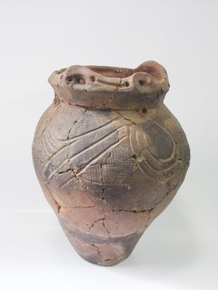 球状の胴体を持つ深鉢形で、口縁部から底部にかけてひび割れや破損箇所があり、口縁部には装飾が施され、胴部には複数の曲線文様と細かな縄目文様が施されている土器の写真