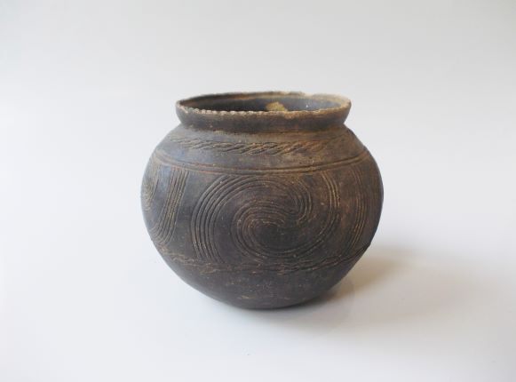 球状の胴体を持つ壺で、胴体には大きな渦巻き模様が複数施され、全体的に黒く変色した土器の写真