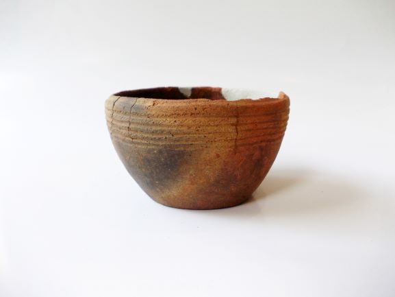 全体的にひび割れや欠けがある茶色の鉢形で、上部の縁には複数の横線が刻まれた土器の写真