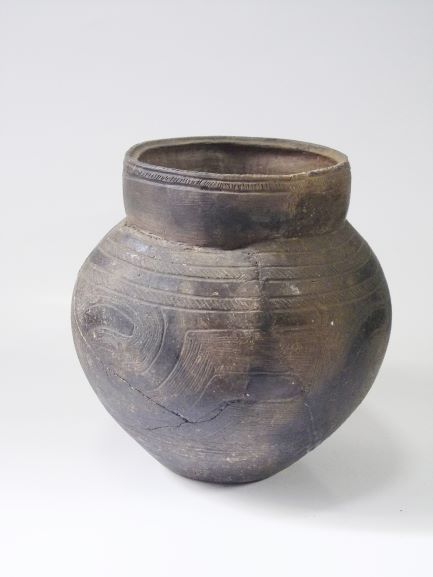 ずんぐりとした胴体と垂直に立ち上がった広い口頸部を持つ暗褐色の土器で、胴体の上部に波線のような複雑な模様が刻まれている注口土器の写真