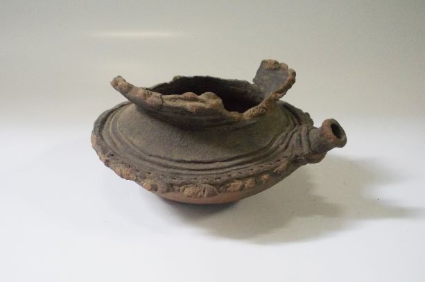 浅い椀状の底部に平たい蓋のような部分が一体成成形され、上部は大きく口が開いている土器で、中央部分の縁には真美打つような装飾があり、側面に注口がある注口土器の写真