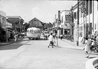 舗装された道路をバスが走っており、周りには多くの店舗や人々が行き交う、昭和時代に撮影されたであろう街中の風景を捉えたモノクロ写真