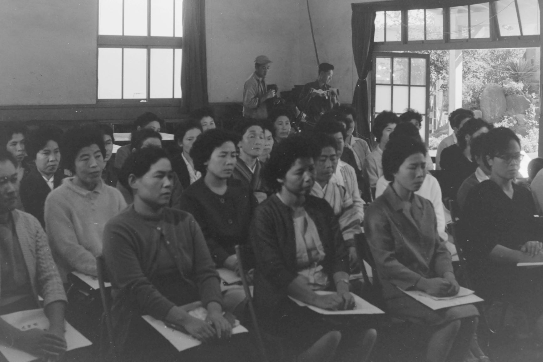 机に向かい、真剣な表情で話を聞く大勢の女性たちが写し出されており、後方にはカメラを構えた男性が立っている、昭和時代の講習会や集会を捉えたモノクロ写真