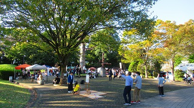 公園の広場で、人々が多数のテントや木のオブジェの周りに集い、イベントを楽しむ様子を写した写真