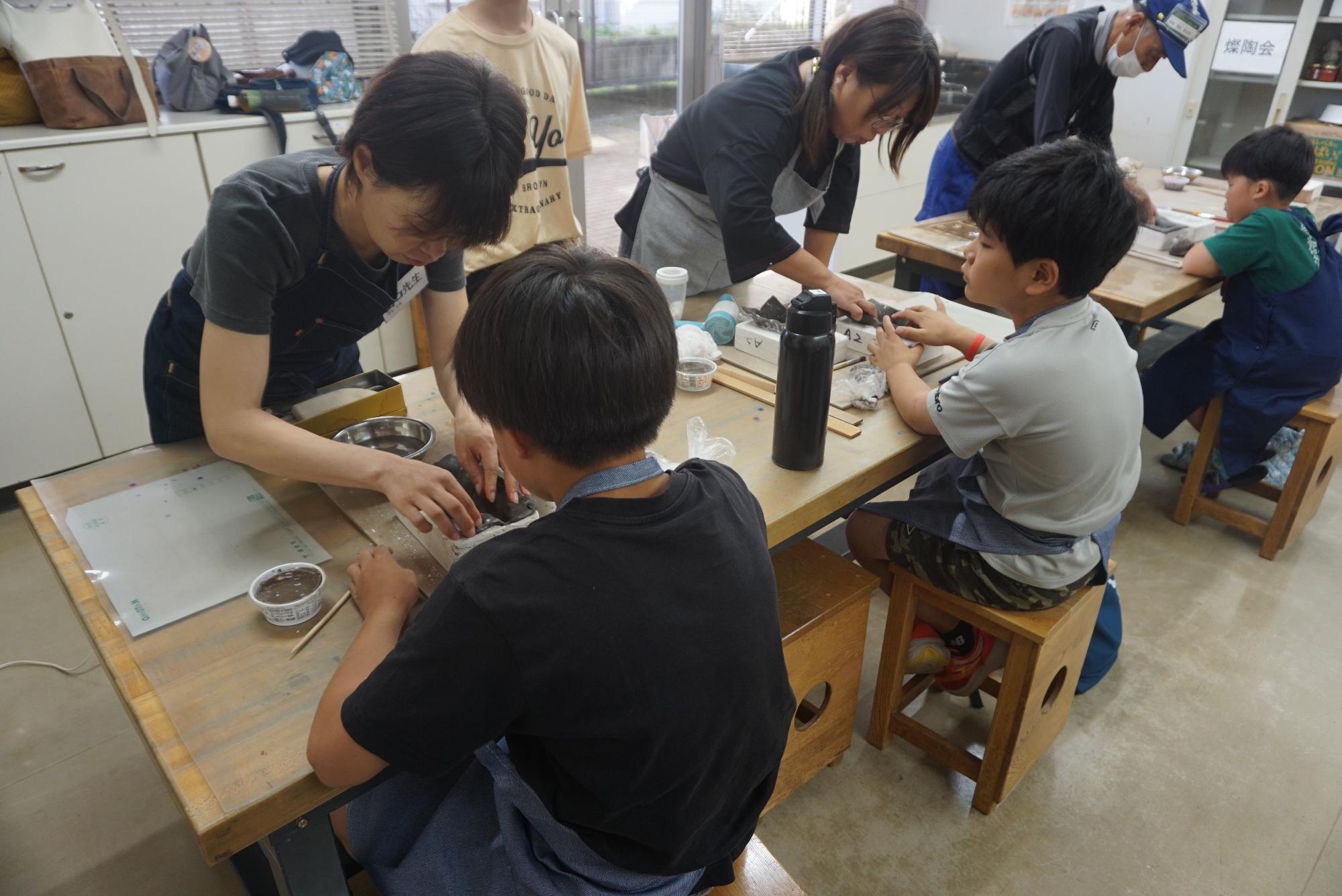 作業台に座った子どもたちが、対面に立った大人とペアになって陶芸をしている様子の写真