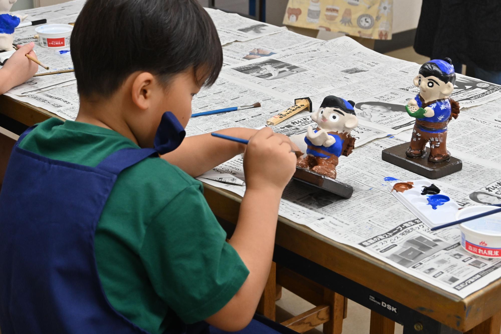 前に置かれた見本を見ながら、新聞紙を広げたテーブルに乗せたミニ金次郎像に着色をしている男の子の写真