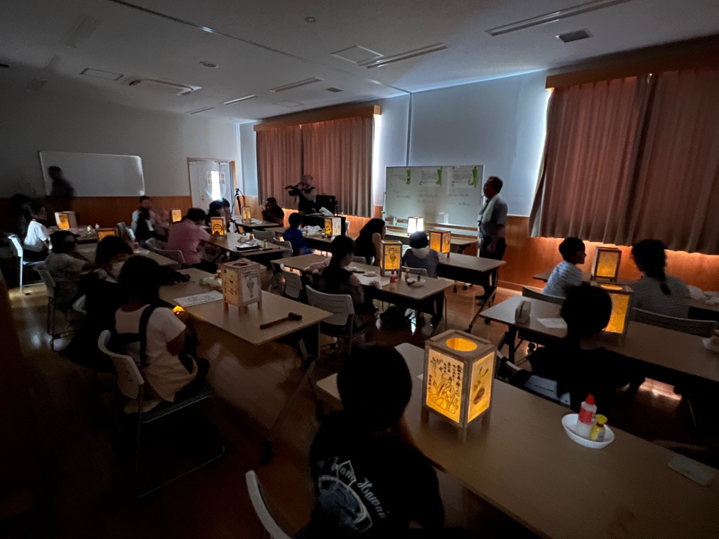 カーテンを閉めて薄暗くした室内で、たくさんの参加者たちが、完成し、明かりをともしたあんどんを前に席に座っている様子の写真