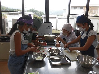 マスクを着用した4人の参加者の子ども達が、ステンレス製の調理台で野菜を切るなどの作業をしている調理実習の様子を捉えた写真