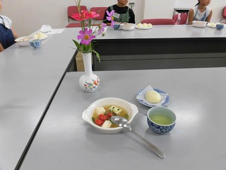 テーブルの上に、花瓶に生けられたコスモスとジニア、ミニトマトと豆腐が入ったスープ、饅頭のような白い菓子、湯呑みに入った緑茶が置かれており、奥には調理実習に参加した子どもたちが着席している様子の写真