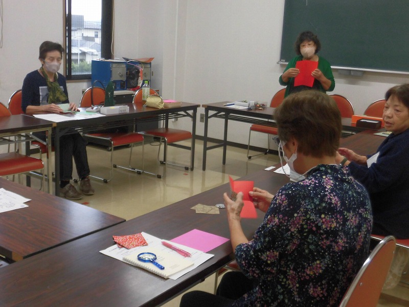 白い壁と黒板のある室内で、数人の女性たちがマスクを着用して机に向かい、手前中央の女性は赤い折り紙を折り進めており、奥の指導者らしき女性も赤い紙を持って説明している様子を捉えた写真