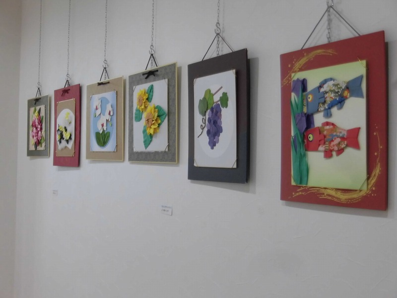 白い壁に、チェーンで吊り下げられた大小さまざまな額縁に入った七つの作品が展示されており、花やブドウ、和柄の鯉のぼりなどが立体的に表現された折り紙の作品が並ぶ写真