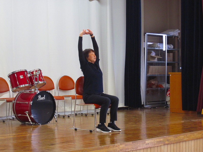 舞台の上で、女性が椅子に座って両手を真上に伸ばすストレッチのような運動を披露している写真