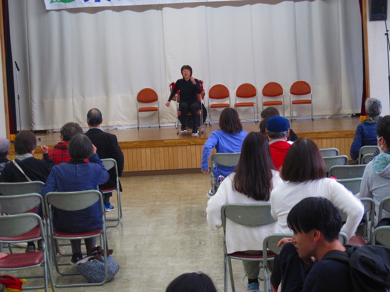 舞台の上で、女性が椅子に座って両手を前に出し、観客席に座る人々に向けて説明や指導をしている様子の写真