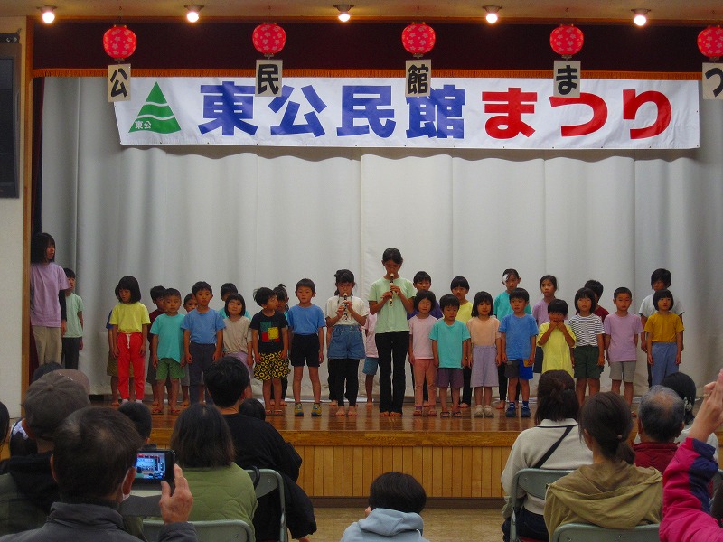 「東公民館まつり」と書かれた横断幕の下、色とりどりの服を着た多くの子供たちが横一列に並び、中央に立つ子供がリコーダーに合わせて発表している様子の写真