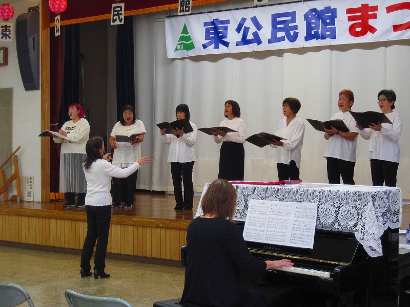 「公民館まつり」と書かれた横断幕が掲げられた舞台の上で、白と黒の衣装を着用した7人のコーラスグループが、ピアノの演奏に合わせて歌を披露している写真