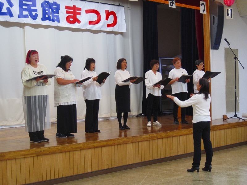 「公民館まつり」と書かれた横断幕が掲げられた舞台の上で、白と黒の衣装を着用した7人のコーラスグループが、指揮者を見ながら歌を披露している写真