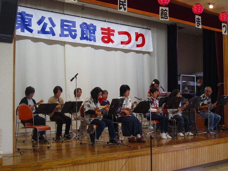 「東公民館まつり」と書かれた横断幕の舞台の上で、楽譜を広げ、椅子に座ってウクレレの演奏をしている写真