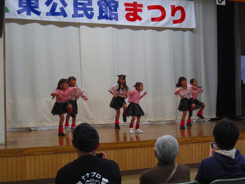 舞台の上で、ピンクの衣装を着用した小学生くらいの子供達が、足上げてダンスを発表している写真